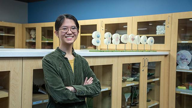 生物学导师凯特琳·赛站在一间科学实验室的房间里，周围的橱柜里放着科学教学工具. 在她的右边是一个展示细胞如何分裂的显示器.  